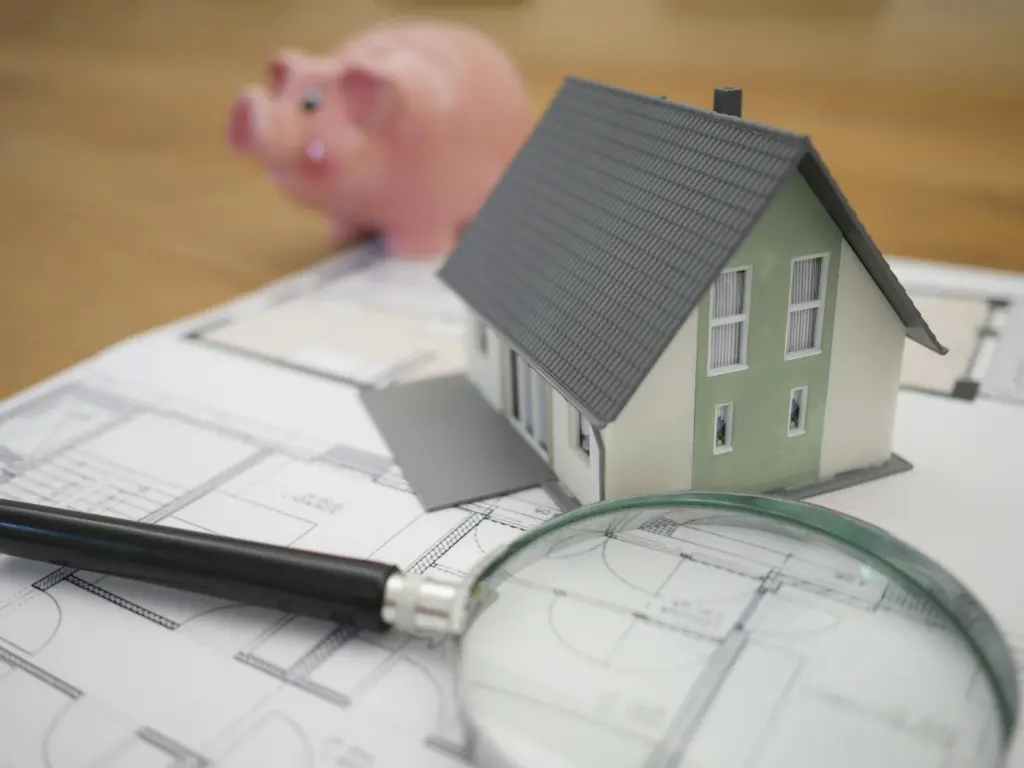 Immobilienbewertung: Aufnahme eines Hausmodells mit einem Sparschwein und einer Lupe auf einem Grundrissplan